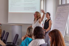 Zwei Referentinnen stehend vor Zuhörer*innen und einem Whiteboard.