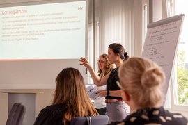 BTZ-Psychologinnen referieren stehend vor einem Whiteboard bei der Agentur für Arbeit. Vor ihnen sitzend Zuhörer*innen an einem Tisch.