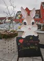 Foto des Aufstellers zum diesjährigen Ostermarkt neben einem geschmückten Osternest. Der Aufsteller selbst besteht aus einem gezeichnete Hasen, der ein Schild mit der Aufschrift Herzlich Willkommen zu Ostern im Frühling hält.
