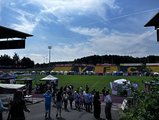 Foto: Zelte mit Beachflags u. Roll-ups rund um den Rasen; viele Besuchende. Dahinter Tribüne mit Werbebannern u. mit gelben/grünen Sitzplätzen, die die Buchstaben VFC abbilden.
