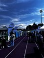 Foto: Messestände mit Roll-ups und Beachflags beiseits der Tartanbahn. Viele Besucher, blauer Himmel mit Wolken. Bäume im Hintergrund, lebendige Stimmung.