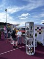 Foto: Person am Infostand auf Tartanbahn, Flaschen in transparenten Aufstellern. Im Hintergrund Stadiontribüne und Rettungswagen, sonnige Stimmung.