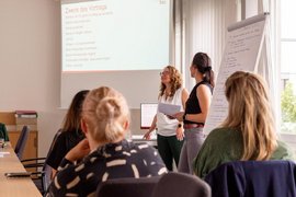 BTZ-Psychologinnen referieren stehend vor einem Whiteboard bei der Agentur für Arbeit. Vor ihnen sitzend Zuhörer*innen an einem Tisch.