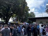 Foto: Menschenmenge auf dem Messegelände unter einem Baum und Vordach, Zelte im Hintergrund, darunter ein gelb-weißes. Sonne scheint bei teils bewölktem Himmel.
