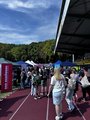 Foto: Menschen auf Tartanbahn erkunden Infostände mit Bannern wie z. B. „DAS HANDWERK“. Stadiontribüne und Bäume im Hintergrund, lebendige Messeatmosphäre.