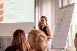 BTZ-Psychologinnen referieren stehend vor einem Whiteboard bei der Agentur für Arbeit. Vor ihnen sitzend Zuhörer*innen an einem Tisch.