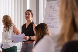 BTZ-Psychologinnen referieren stehend vor einem Whiteboard bei der Agentur für Arbeit. Vor ihnen sitzend Zuhörer*innen an einem Tisch.