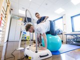 Einblick in die Therapiearbeit der NTRA: Eine Person balanciert mit den Knien auf einem Therapieball, mit en Händen auf ein Gerät gestützt. Hinter der Person steht ein Mitarbeiter der NTRA und unterstützt.
