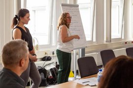 BTZ-Psychologinnen referieren stehend vor einem Whiteboard bei der Agentur für Arbeit. Vor ihnen sitzend Zuhörer*innen an einem Tisch.