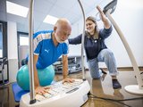 Einblick in die Therapiearbeit der NTRA: Eine Person balanciert mit den Knien auf einem Therapieball, mit en Händen auf ein Gerät gestützt. Neben der Person kniet eine Mitarbeiterin der NTRA und unterstützt.