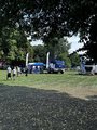 Foto: Messestand THW mit Ausstellenden u. Besuchenden auf einer Wiese, von Bäumen umgeben. Zwei LKWs des THW stehen neben dem blauen Pavillon mit weißen Fahnen.