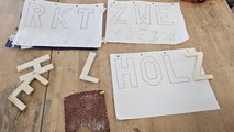 Vorbereitung der Arbeit Foto: Drei Skizzenblätter mit aufgezeichnete Buchstaben sowie vier fertige Holzbuchstaben liegen auf der Werkbank.