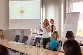 Zwei Referentinnen stehend vor Whiteboard und Zuhörer*innen.