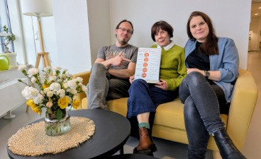 Foto des Team Kundenservice: Drei Personen sitzen auf einer Couch, davor ein runter Tisch mit einem Blumenstrauß darauf. 