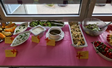 Sommerfest im Beruflichen Trainingszentrum Auf dem Foto ist ein Buffet zu sehen, das auf einem Tisch, mit einer roten Tischdecke, aufgebaut wurde. Dieser steht am Fenster. Zu sehen sind Salate und Saucen.
