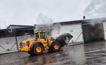 Das Bild zeigt einen gelben Frontlader auf dem ZAK-Gelände in Kaiserslautern. Er hebt eine dampfende Ladung vor einem Gebäude mit weißen Toren. Der Boden ist nass – es hat geregnet. Die Szene wirkt industriell und zeigt aktive Arbeit in der Abfallverwertung.