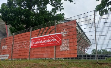 Zu sehen ist ein hoher Metallzaun, an dem ein Banner angebracht ist, das den Weg zur Ausbildungsmesse zeigt.