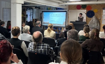 Foto vom Raumende über die Zuhörenden, die in Reihen sitzend zum/zur Redner*in und der Präsentation am Whiteboard blicken. 