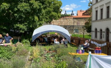 Foto: Gemütliches Sommerfest im Grünen: Menschen sitzen entspannt unter einem weißen Pavillon, umgeben von blühenden Pflanzen und historischer Architektur.