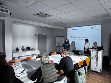 Foto: Personen sitzen in einem Seminarraum und arbeiten an Unterlagen, während eine Präsentation an der Wand gezeigt wird.