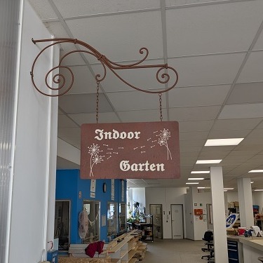 Braunes Schild mit Aufschrift "Indoorgarten", welches an einer gusseisernen Befestigung an der Wand vorm Eingang des Indoorgartens hängt