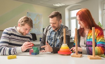 Drei Personen sitzen am Tisch, zwei von ihnen sind mit einem Trainingsmittel aus Holz beschäftigt. Die dritte Person begleitet die Aufgaben.